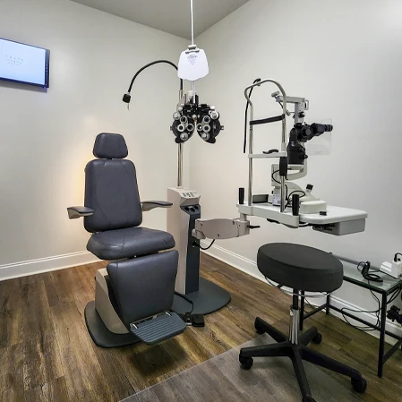 Exam room with ophthalmic chair, phoropter, slit lamp, stool, wall-mounted screen, and wood flooring.