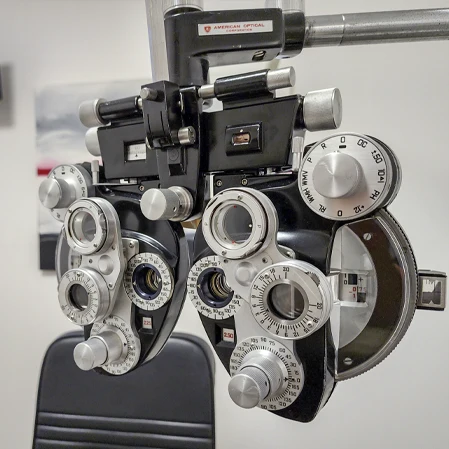 Close-up of a phoropter with multiple dials and lenses mounted in front of an exam chair in a clinic room.